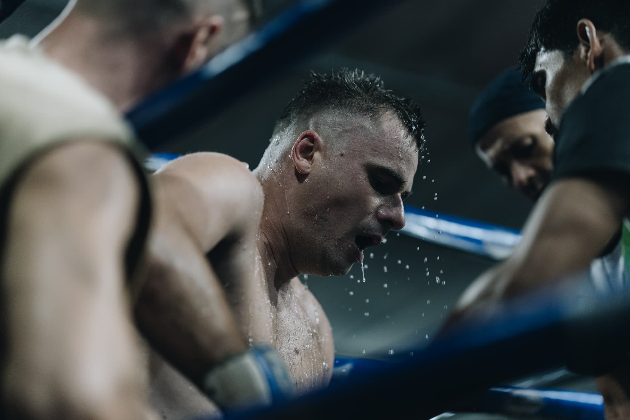 Water drips from a weary, sweat-soaked boxer as his corner team provides support mid-fight.