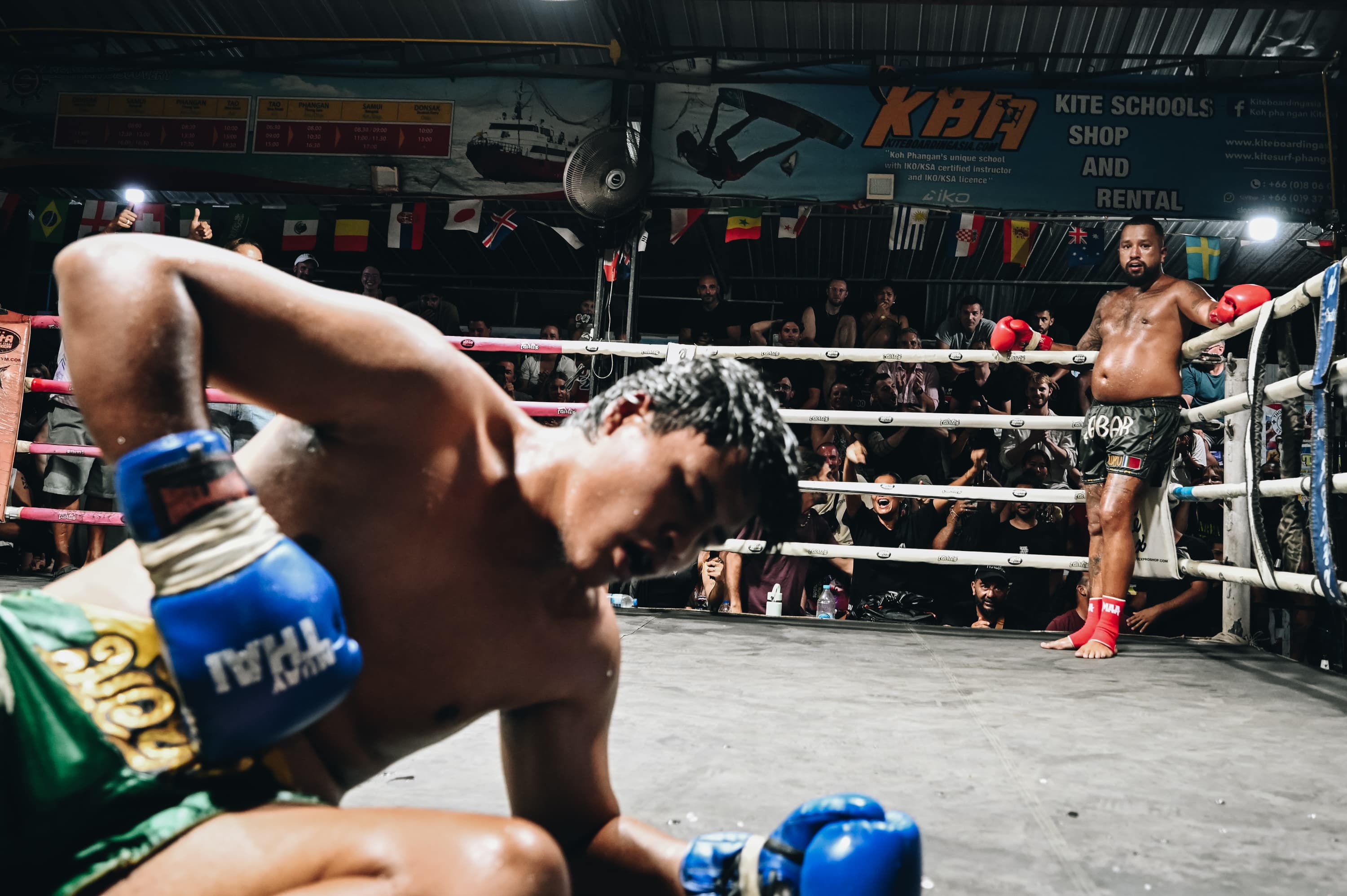 Muay Thai fighter on the canvas after a knockdown while his opponent watches nearby.