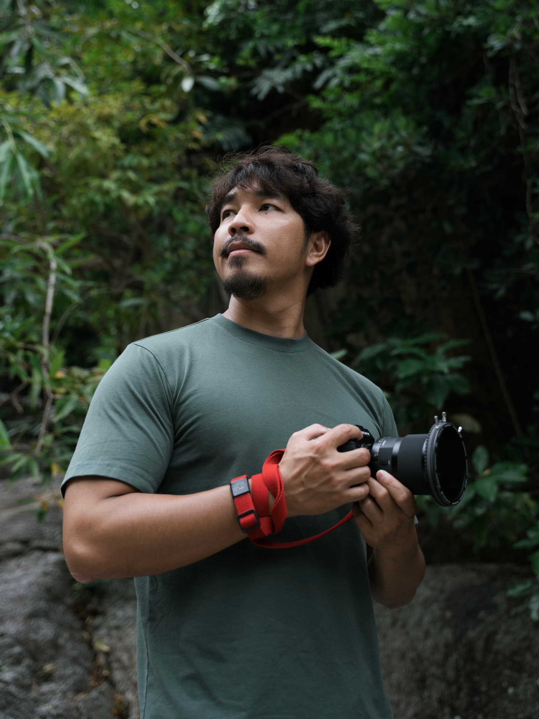 Bearded man in a green shirt holding a camera, looking up amidst dense forest foliage.