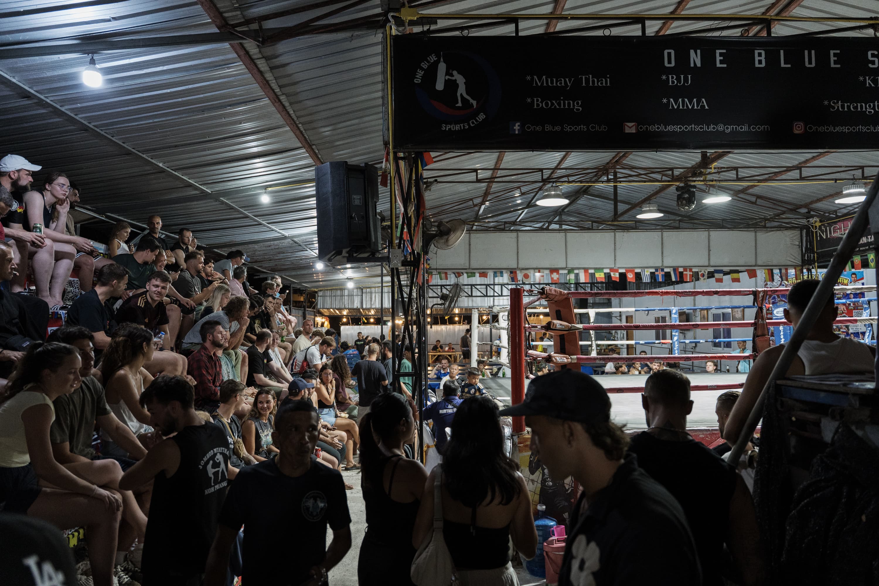 Crowd on bleachers overlooking a boxing ring in a dimly lit indoor sports club.