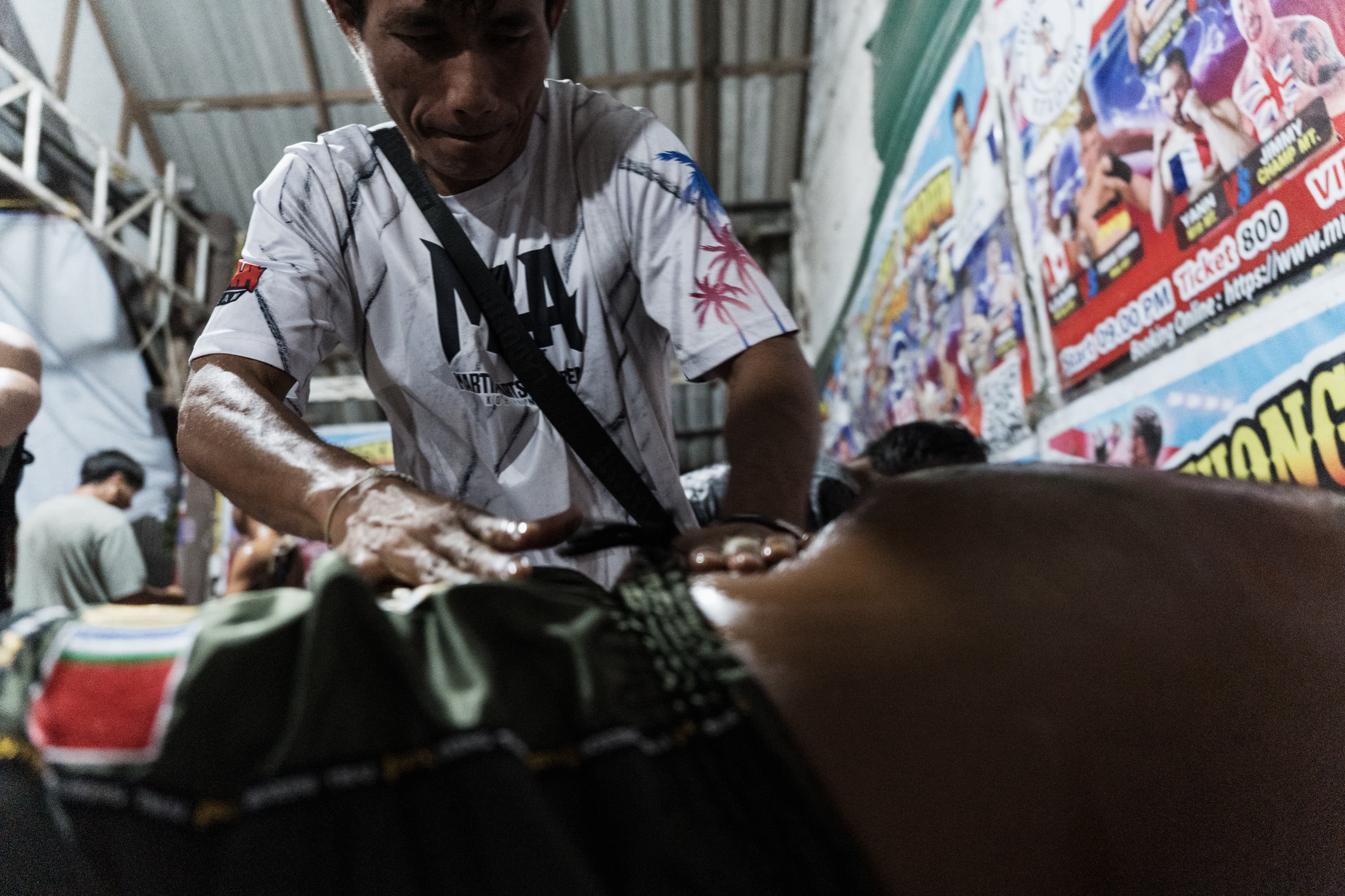 Man applies oil to a Muay Thai fighter's back in a gym with fight posters.
