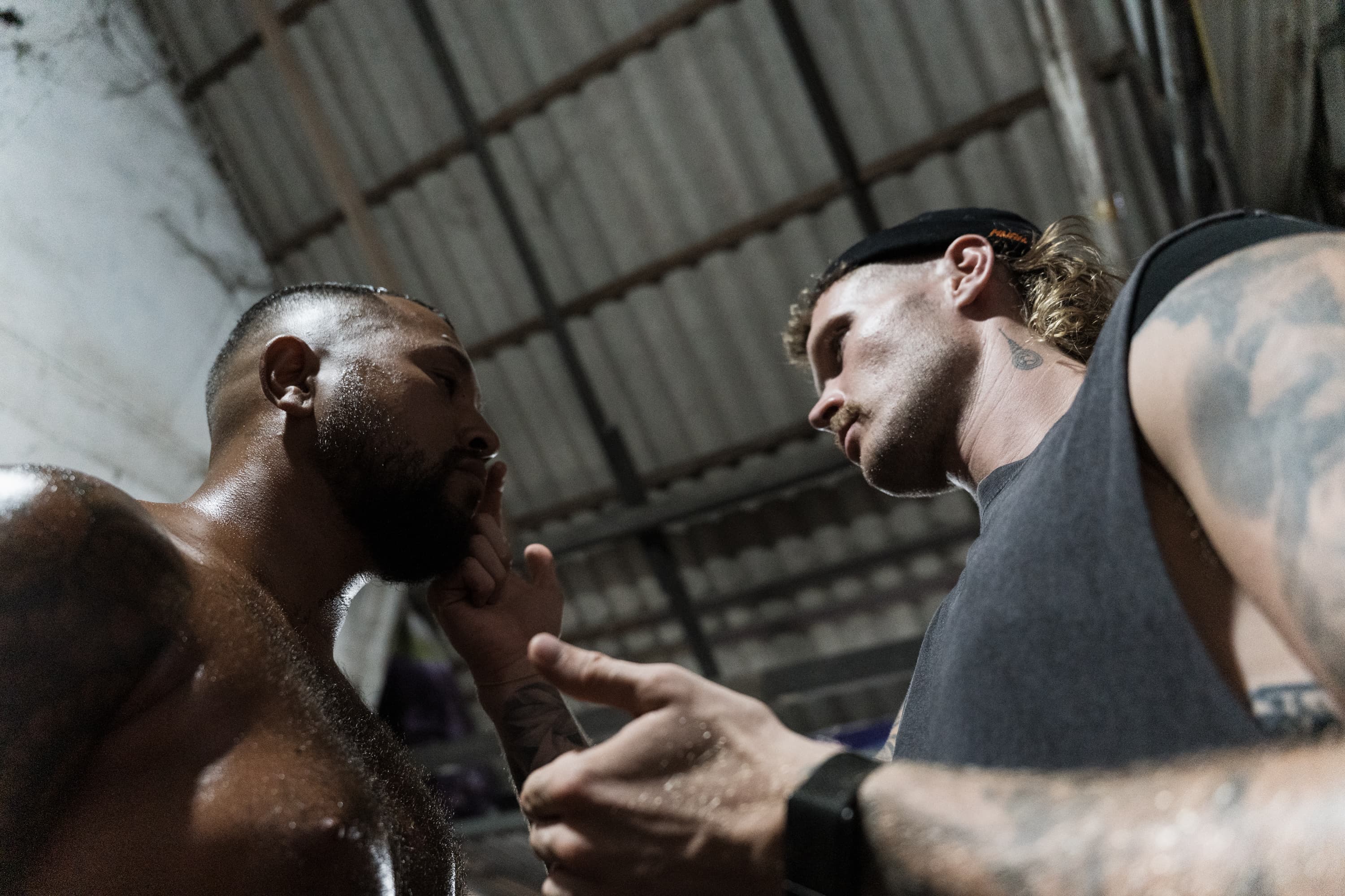 Two tattooed men engage in a tense, close-up confrontation inside a dimly lit gym.