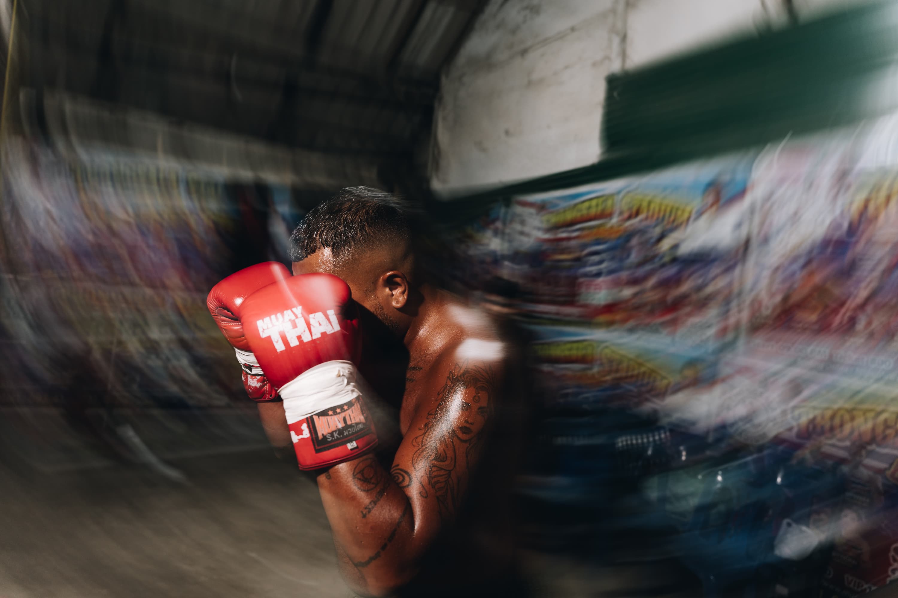 Tattooed fighter in red Muay Thai gloves guarding his face amidst streaks of motion blur.
