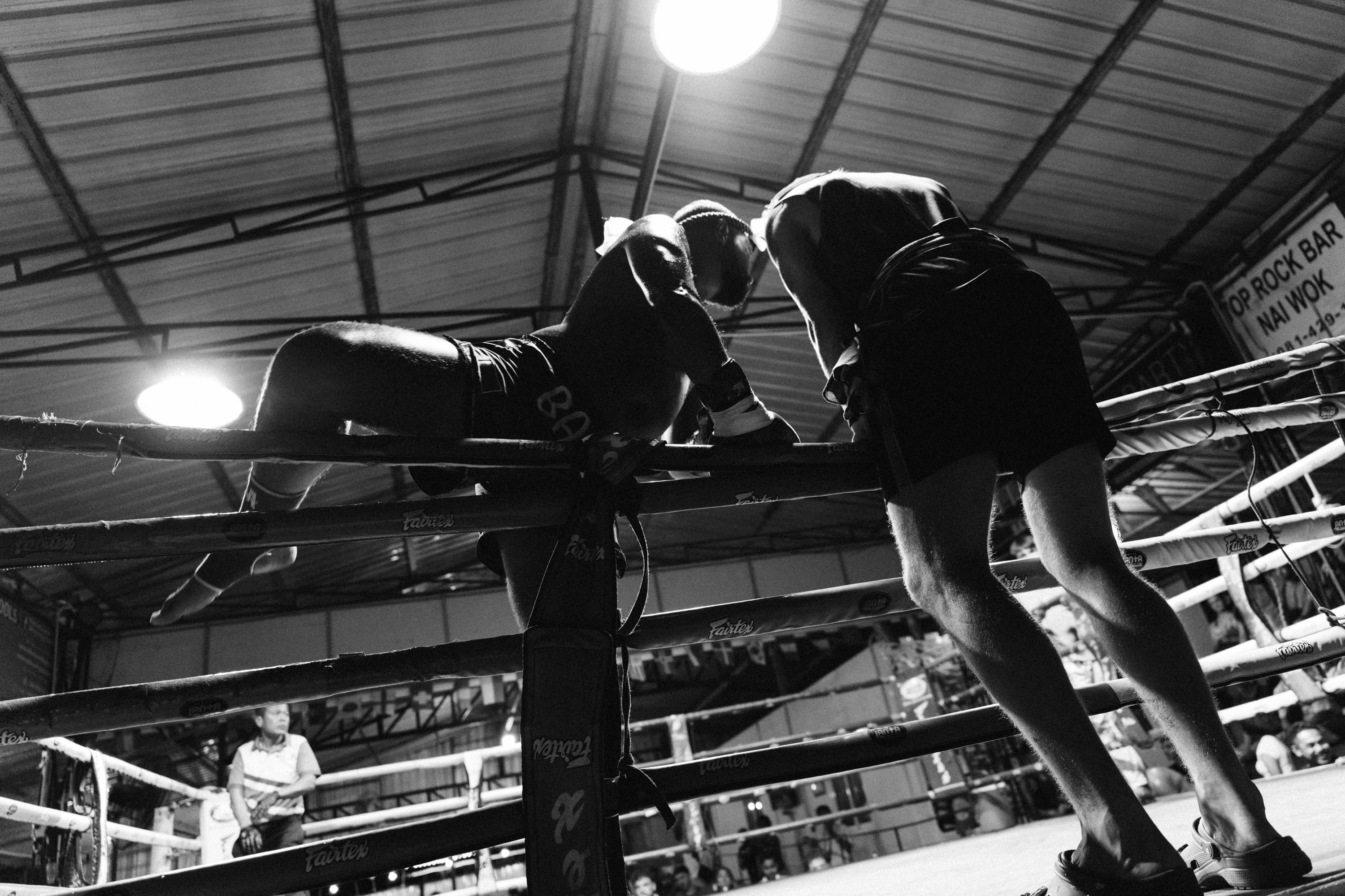 Low-angle black and white shot of a fighter climbing into a bright Muay Thai ring.
