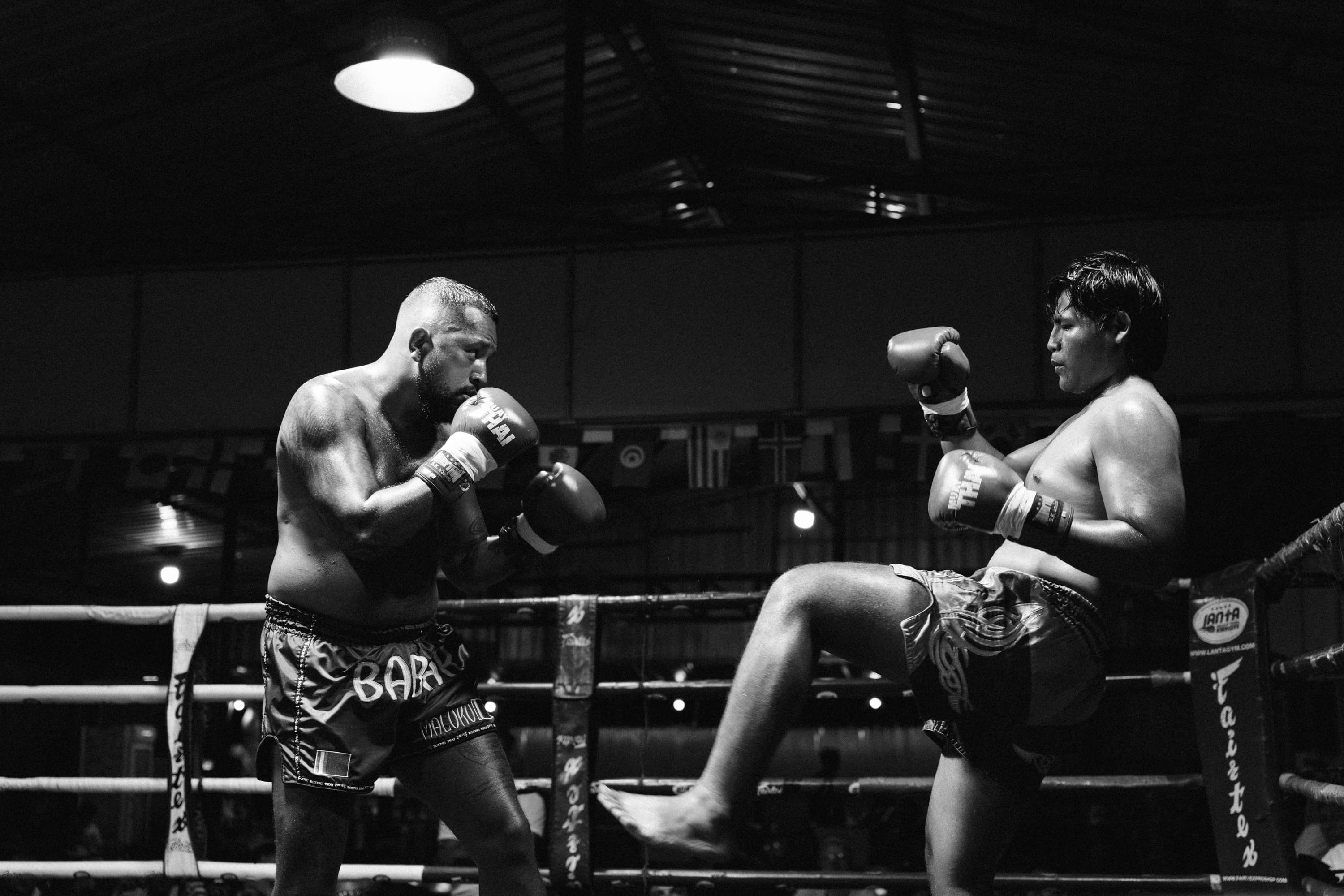 Two Muay Thai fighters sparring in a dimly lit ring, one throwing a powerful kick.