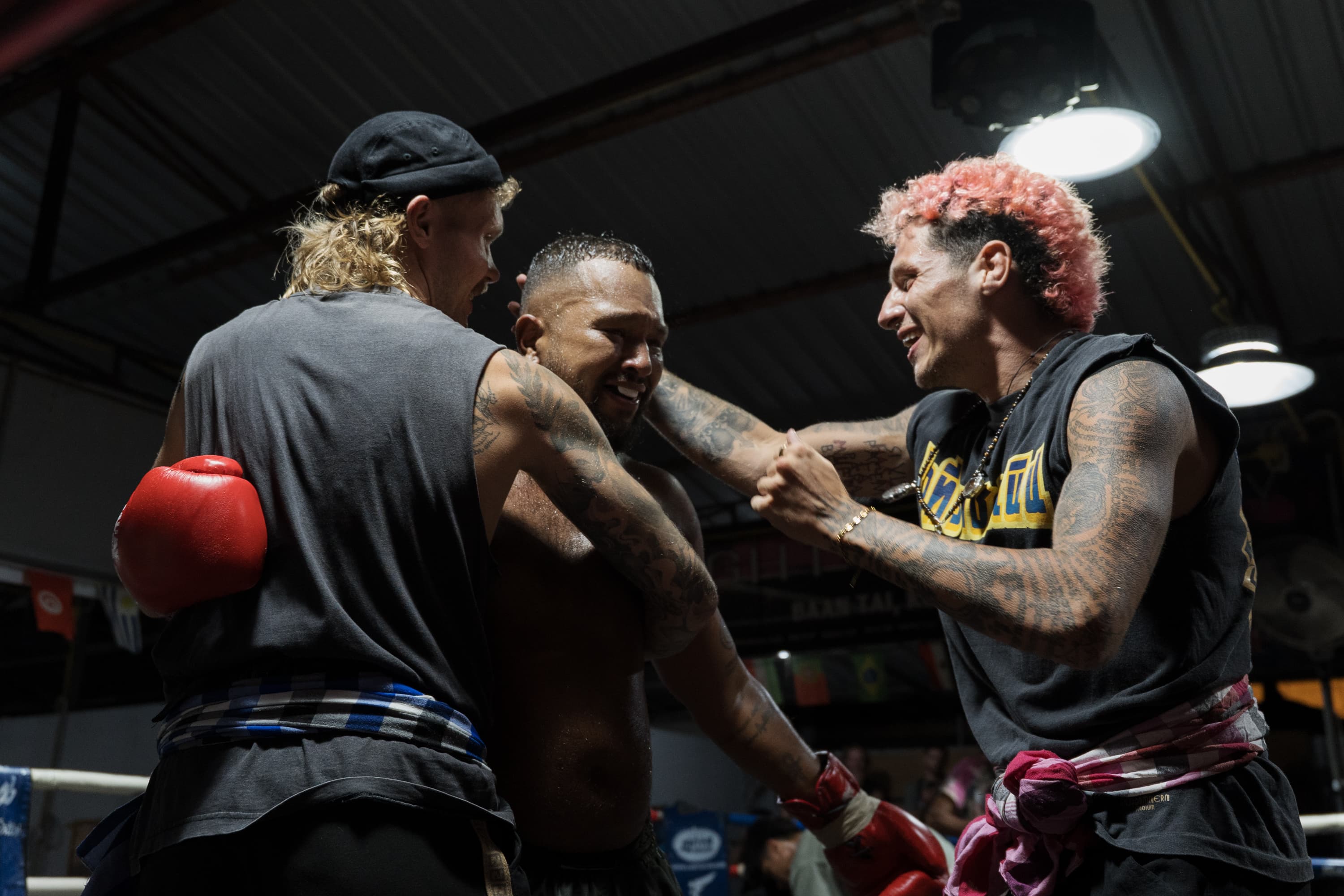 Two tattooed men embrace and congratulate a sweaty boxer inside a dimly lit boxing ring.
