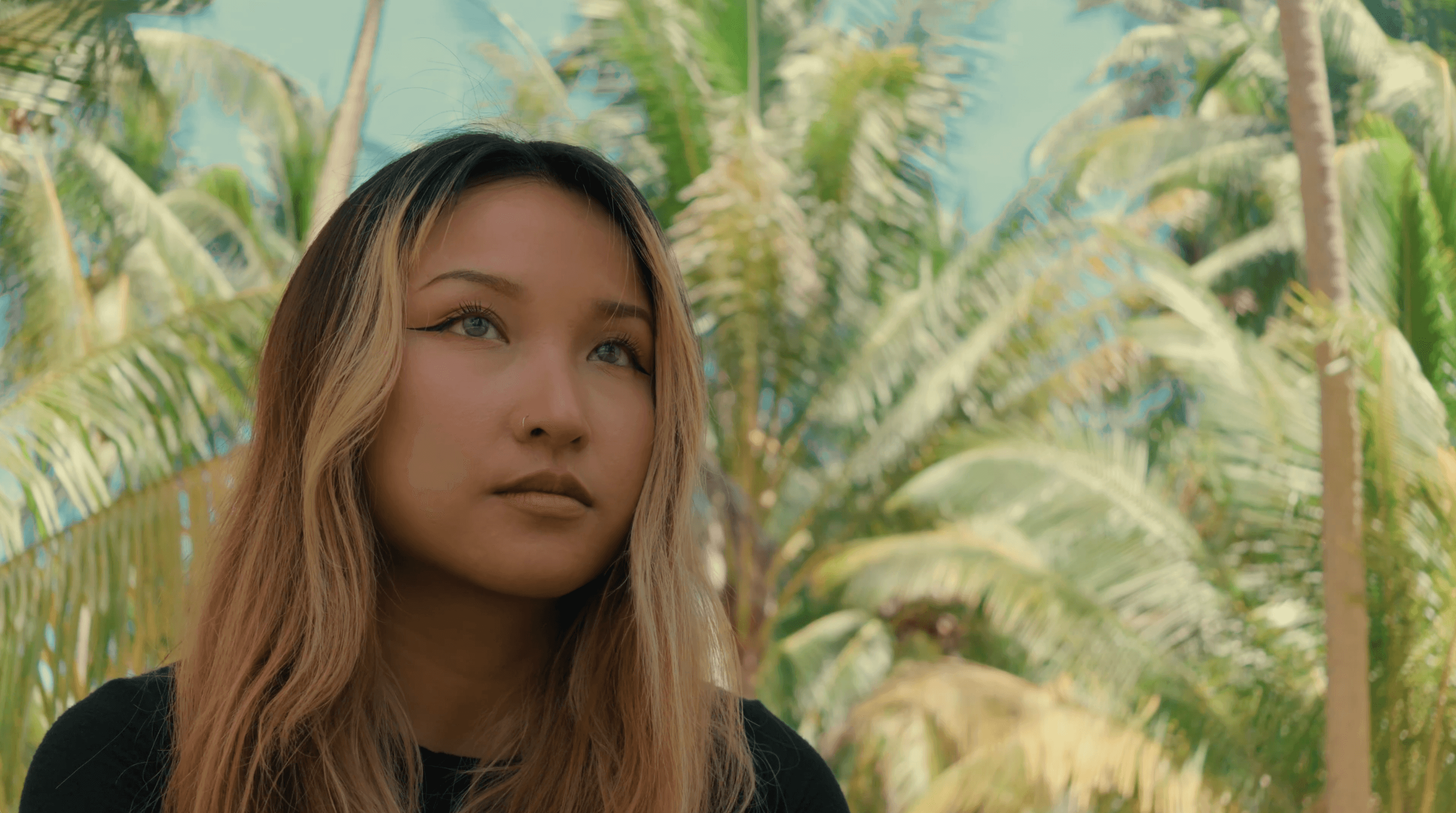 Pensive woman with blonde highlights looking away against a sunny background of tropical palm trees.