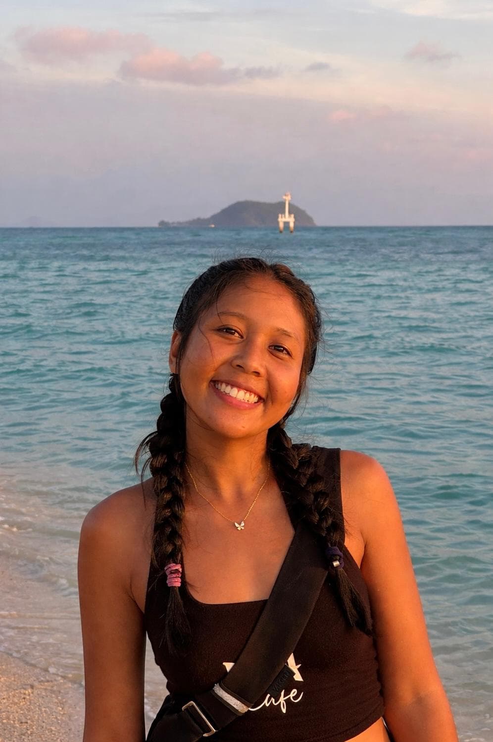 Smiling woman with braided hair on a beach at sunset with turquoise water and island.